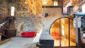 Habitación de paredes de piedra con un sofá rojo, una puerta de cristal circular que da a un espacio soleado y una escalera con vigas de madera.