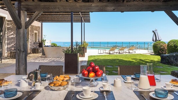 Un espacio de comedor al aire libre con una mesa preparada para el desayuno, con vista a una piscina y al océano.