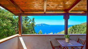 Een overdekt balkon met een tafel en stoelen, met uitzicht op een schilderachtig zeegezicht en een bergachtig landschap.