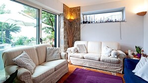 A living room with two sofas, a stone accent wall, a window with a view of palm trees, and a small plant. - Villa Gesine with private pool and sea views (Massa Lubrense)
