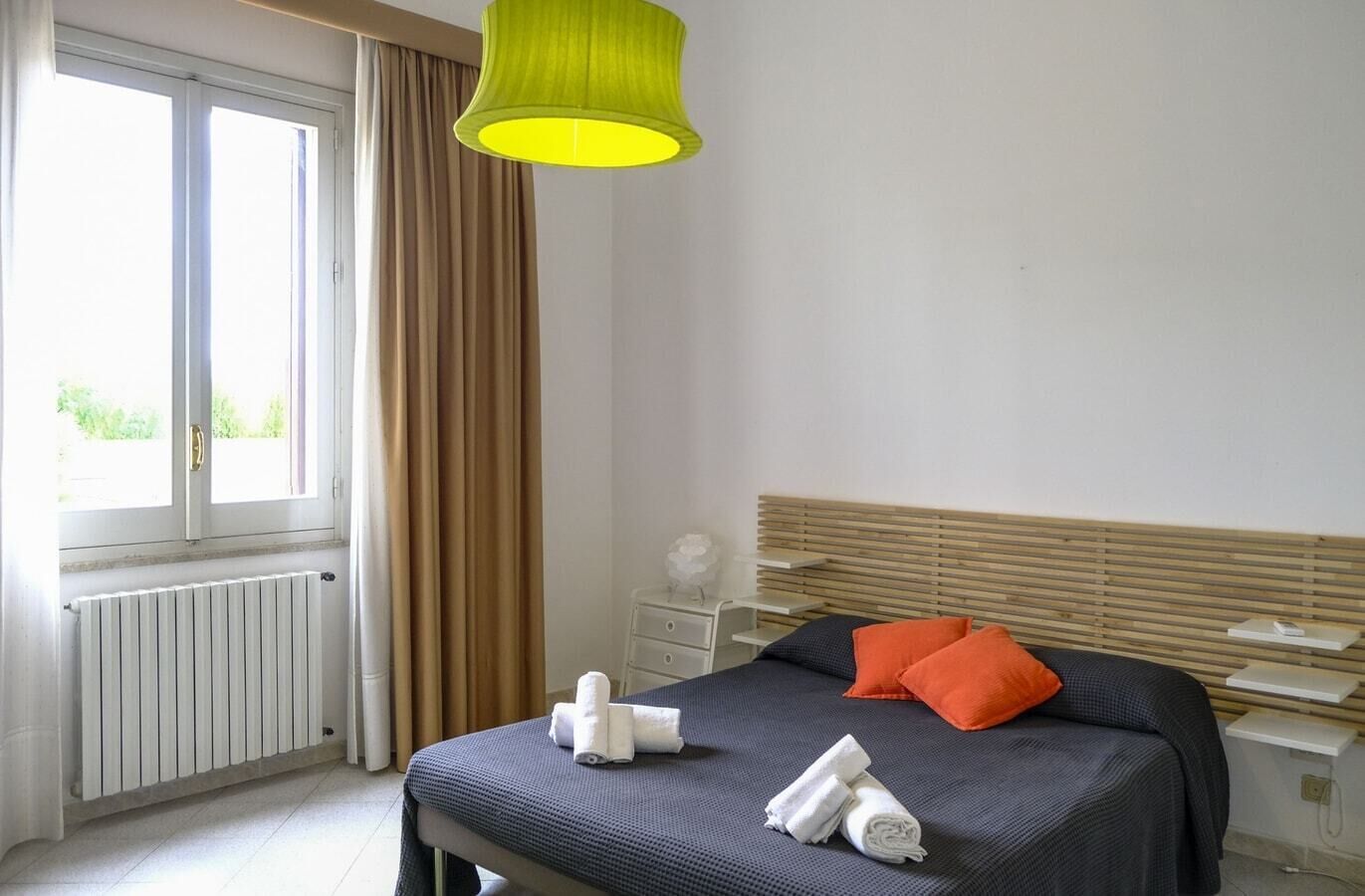 Habitación de hotel con cama, mesita de noche, lámpara, ventana con cortinas y radiador.