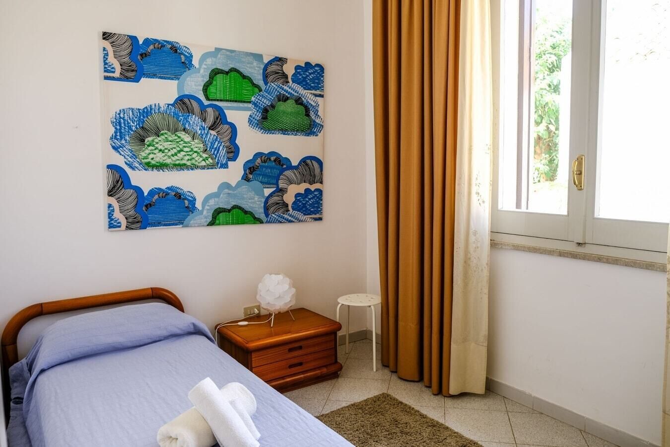 A bedroom with a bed, a nightstand, a wall art piece, a window with curtains, and a view of greenery outside.