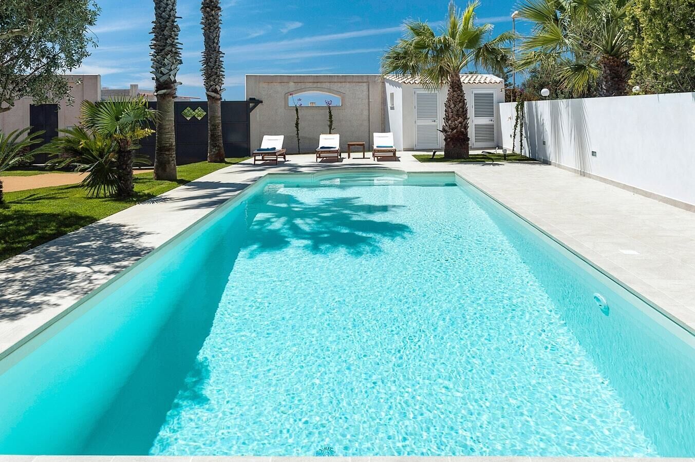 A rectangular swimming pool with clear blue water, surrounded by a white wall and palm trees. There are two lounge chairs with blue cushions on the poolside.