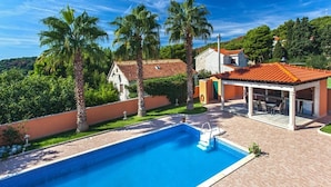 A swimming pool with a patio area, surrounded by palm trees and a house with a red-tiled roof. - Villa Venera - Coastal Villa with pool in Sumartin (Sumartin)