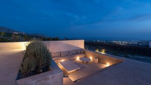 Eine Dachterrasse mit einer Couch und einem runden Tisch, umgeben von Pflanzen und beleuchtet durch stimmungsvolle Beleuchtung.