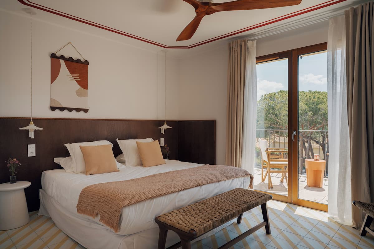 Un dormitorio con una cama grande, cabecera de madera, un banco, ventilador de techo y vistas al exterior.