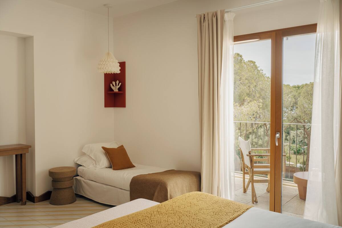 Un dormitorio con cama, mesita de noche, una silla y vistas a un balcón con mesa y sillón.