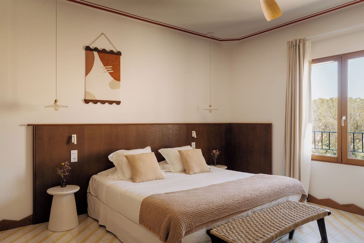 Un dormitorio con una cama grande, un cabecero de madera, una mesita de noche con un florero y una ventana con vistas.