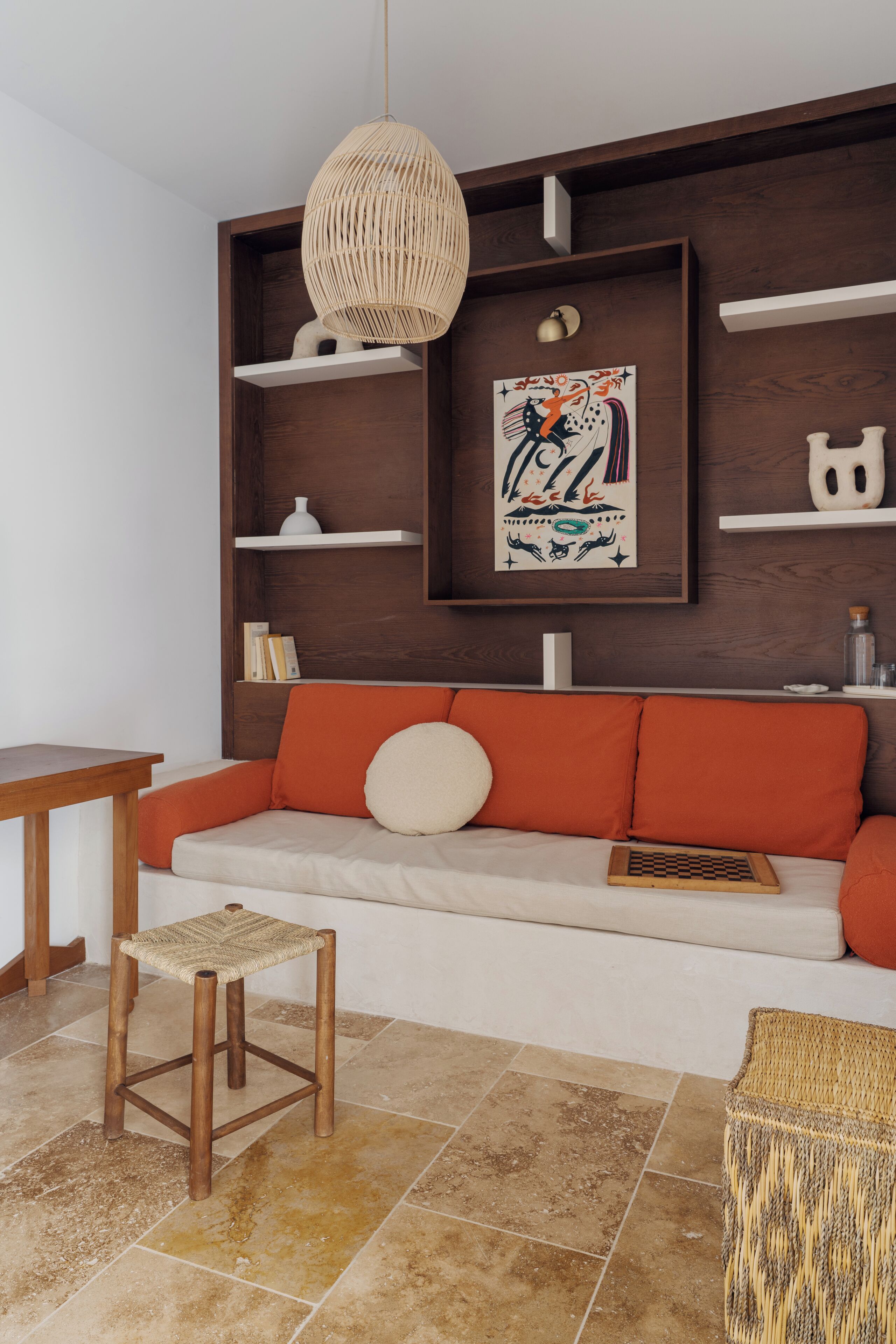 A modern living room with a wooden bench, a woven stool, and a sofa with orange cushions.