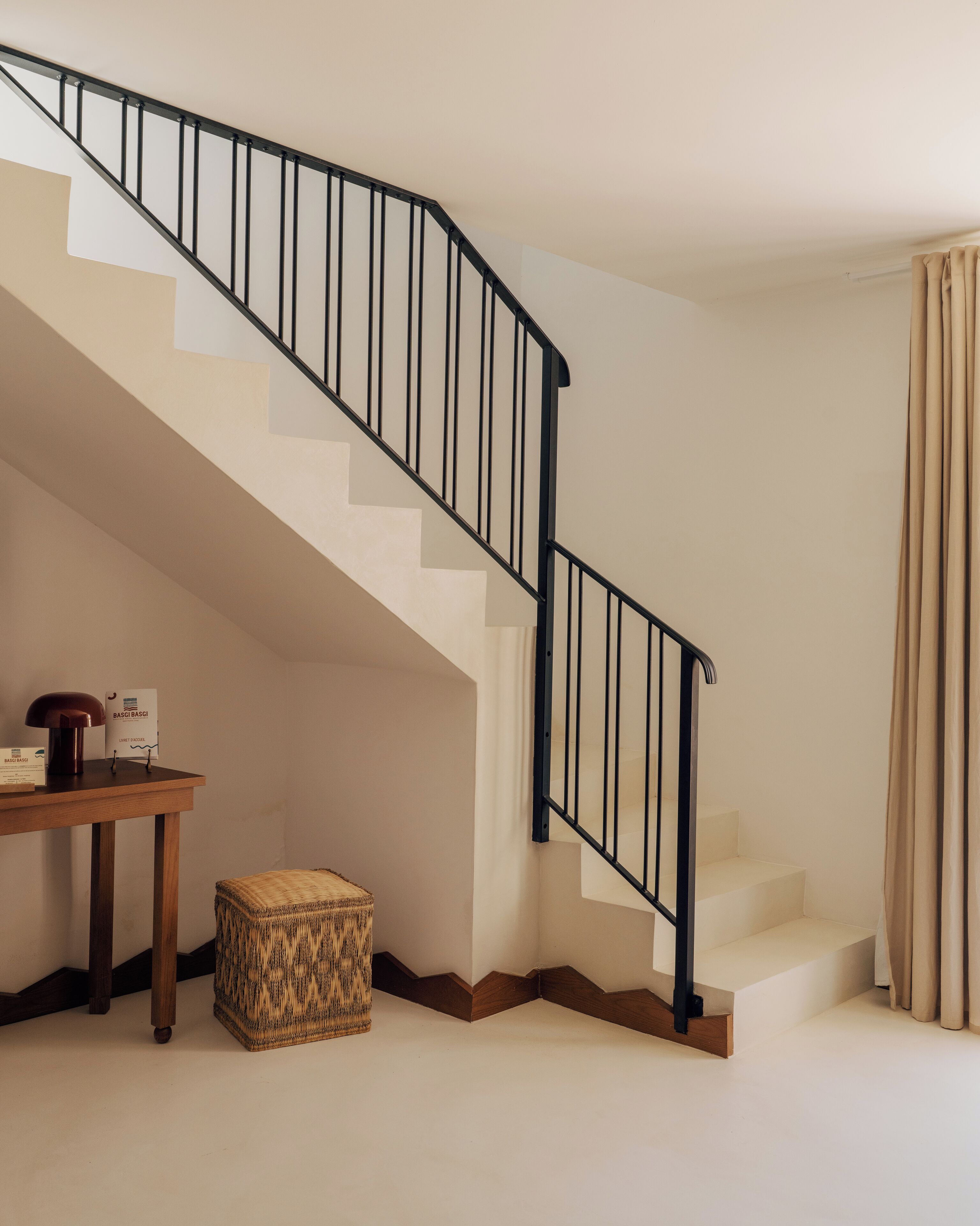 Una escalera con barandal negro, una mesa de madera con una lámpara y una caja, y un reposapiés tejido.