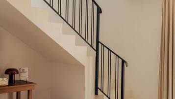 Una escalera con barandal negro, una mesa de madera con una lámpara y una caja, y un reposapiés tejido.