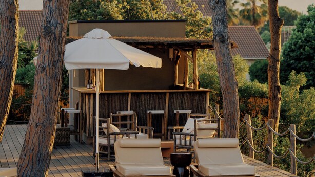 A wooden deck with lounge chairs and a pool, surrounded by trees and a building with a thatched roof.