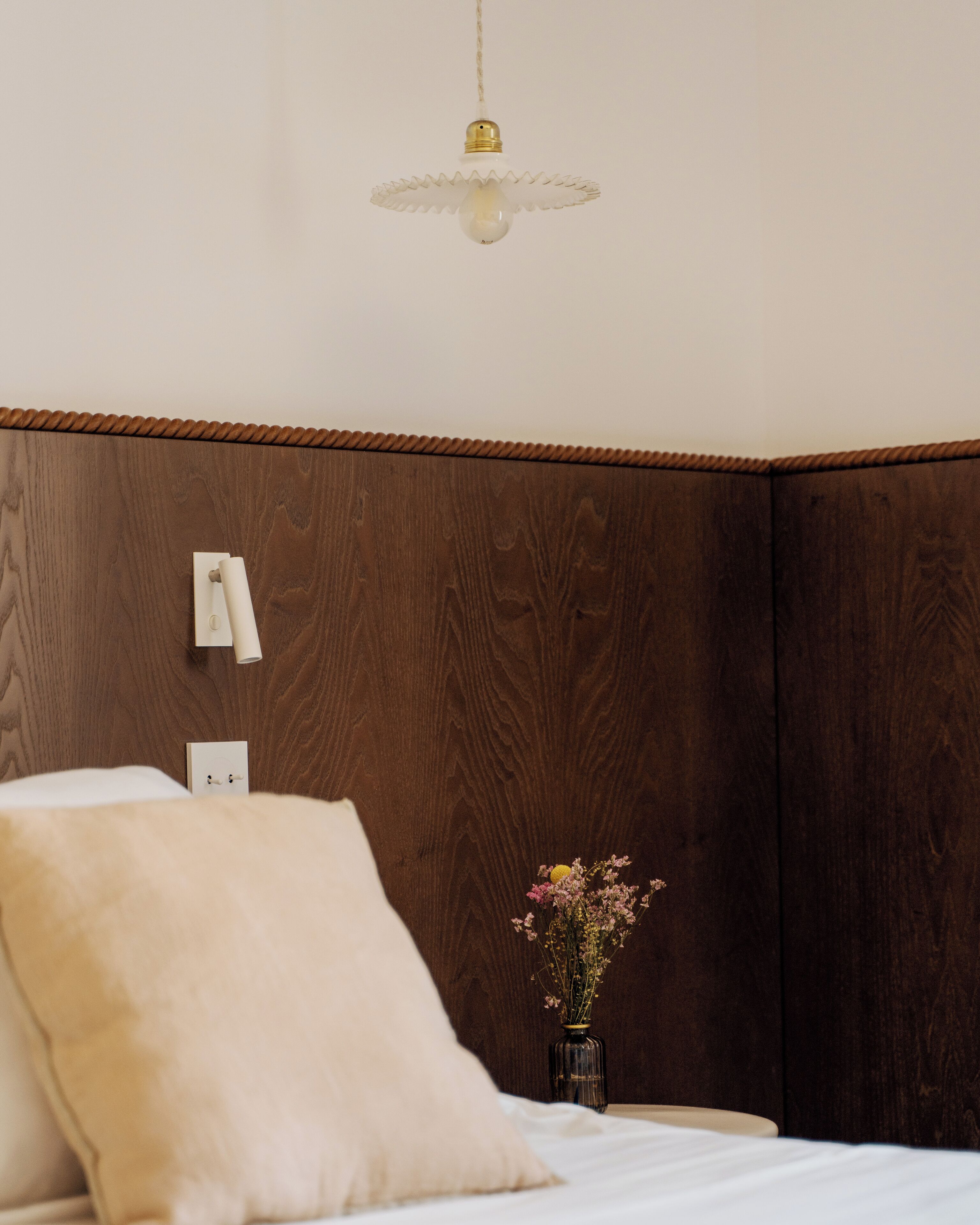 A bedroom with a wooden headboard, a bedside table with a vase of flowers, and a hanging light fixture.