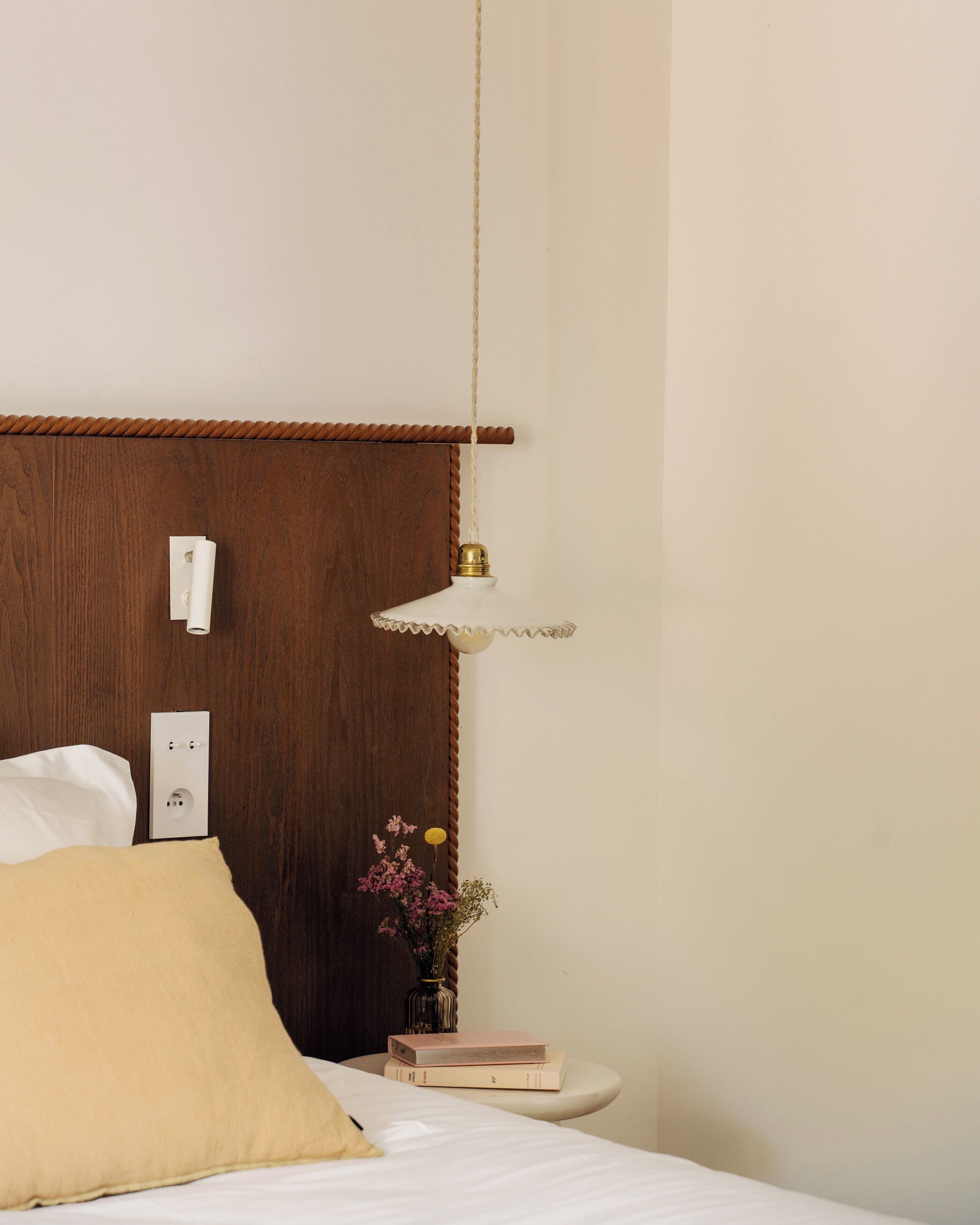 A neatly made bed with a pillow, a bedside table with a book and a vase of flowers, and a wall-mounted light fixture.
