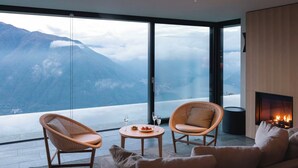 A modern living room with a large glass wall offering a mountain view. There are two wicker chairs, a small round table with a glass of wine, and a sofa. A fireplace is visible in the background. - Villa Oltre Le Nuvole - Luxury villa with lake views (Pigra)