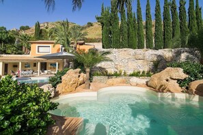 A pool area with a stone wall, palm trees, and a building with a tiled roof. - Villa Michelangelo - Stunning family villa (Agrigento)