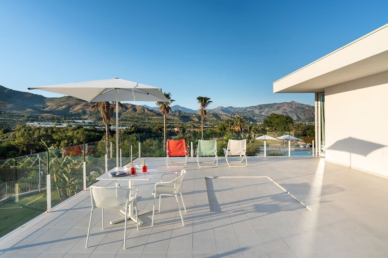 A modern outdoor terrace with a clear view of mountains, equipped with white chairs, tables, and umbrellas.