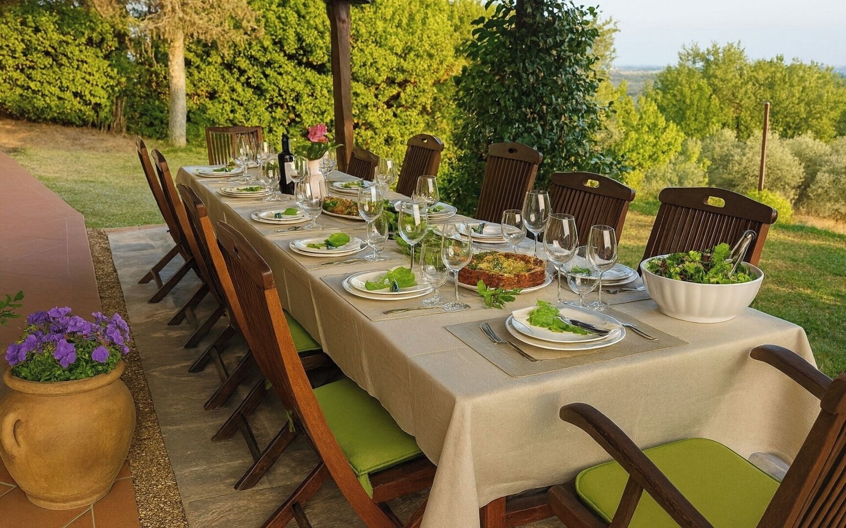 An outdoor dining table set with plates, glasses, and a bowl of salad, surrounded by chairs.