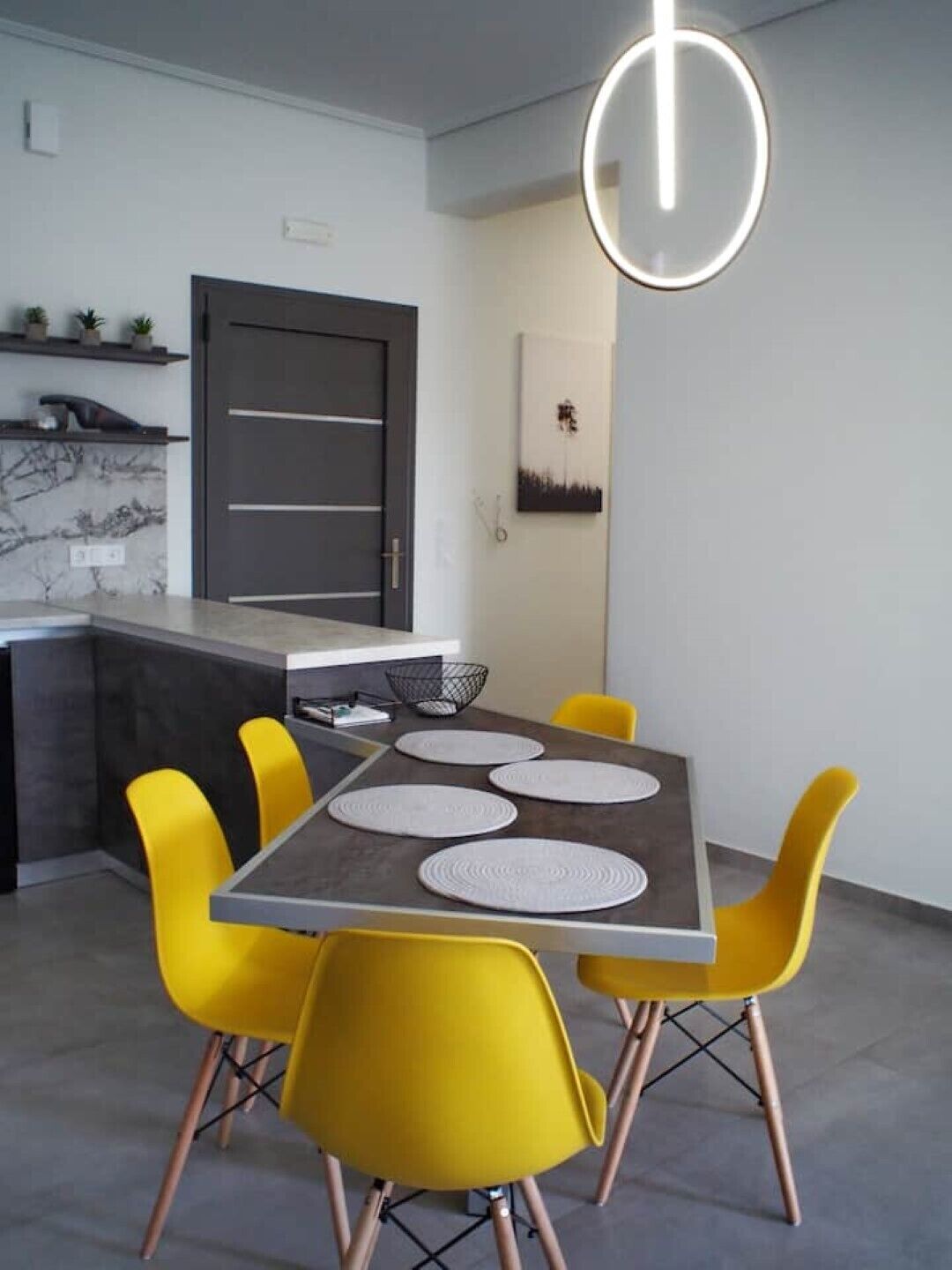 Une cuisine moderne avec une porte gris foncé, un plan de travail en marbre et un coin repas comprenant une table grise et des chaises jaunes.