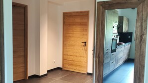 Een gang met een houten deur, een betegelde vloer en een keuken met kastjes en een koelkast.