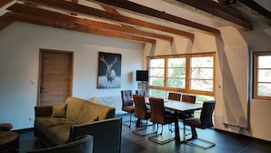 Een kamer met een houten plafond, een eettafel met stoelen, een bank, een radiator en een raam met gordijnen.