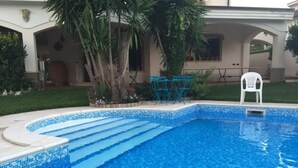 Une piscine dont le bord est recouvert de carrelage, entourée d’une pelouse et de palmiers.