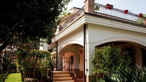 Une maison à deux étages dotée d’un porche abrité, d’un escalier menant à l’entrée et d’un jardin agrémenté de plantes en pot et d’une pelouse.