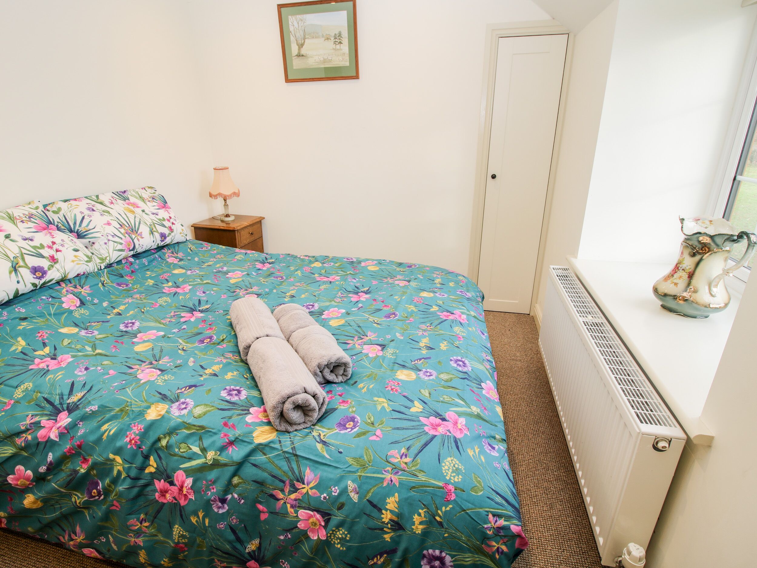 A bedroom with a floral bedspread, a rolled towel, a radiator, and a picture on the wall.