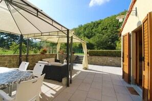 En udendørs terrasse med overdækket siddeområde, stenvægge og udsigt til en skovklædt bakke.