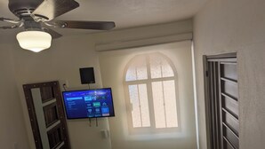 A room with a ceiling fan, a mounted television displaying a game interface, a window with a view, and a door with a metal grill. - Moderna, Cómoda Suite Nube en Tepotzotlán centro (Tepotzotlán)