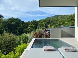 Une piscine extérieure moderne, dotée de chaises longues et de serviettes, entourée d’une végétation luxuriante et offrant une vue sur une colline.
