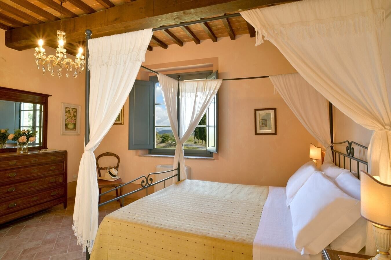 A bedroom with a bed, a wooden dresser, a chandelier, and a window with curtains.
