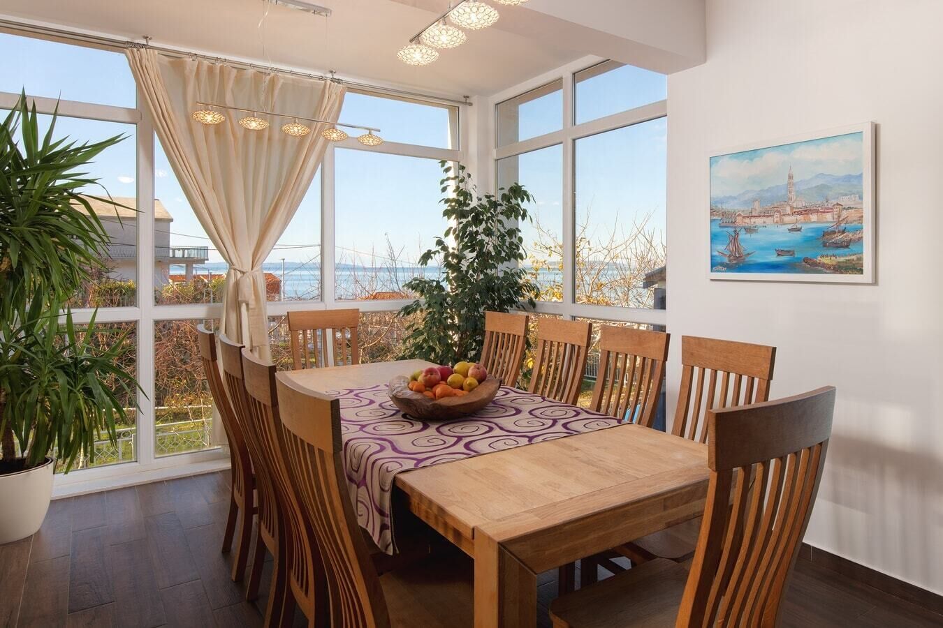 Un'area pranzo con tavolo e sedie in legno, una ciotola di frutta e vista mare attraverso ampie finestre.