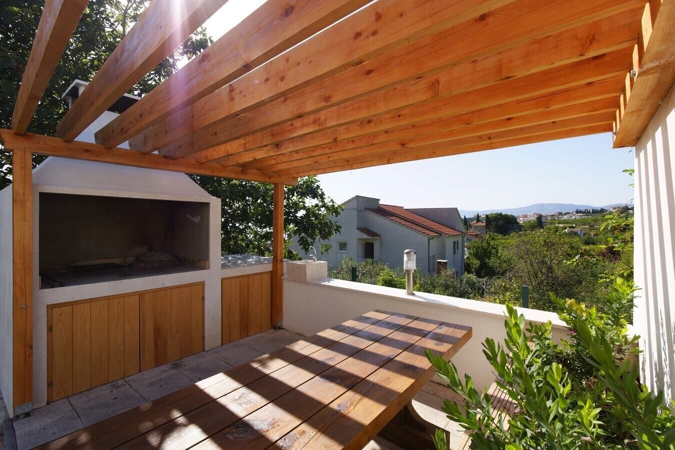 Una pérgola de madera con zona de barbacoa y una mesa, con vistas a una zona residencial con casas y vegetación.