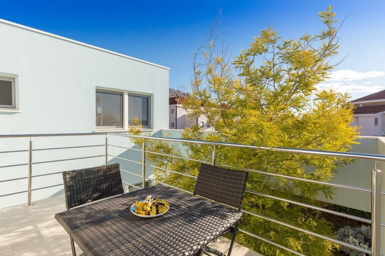 Un balcon extérieur avec une table dressée pour deux personnes, entouré de rampes en métal, et un arbre aux feuilles vertes en arrière-plan.