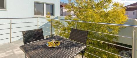 Un balcon extérieur avec une table dressée pour deux personnes, entouré de rampes en métal, et un arbre aux feuilles vertes en arrière-plan.
