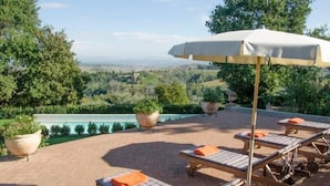 A poolside area with lounge chairs and an umbrella, surrounded by greenery and a view of the landscape. - Villa Elara - Tuscan Countryside Villa withPool (Montaione)