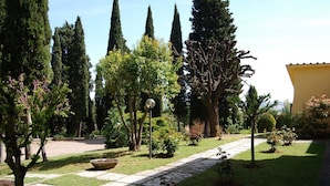 A garden with a stone pathway, various trees, and a yellow building in the background. - Villa Elettra - Private villa with pool in Umbria (Castiglione del Lago)