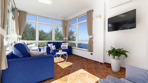 A spacious living area with a blue sofa set, a small white table, and a striped sofa. Large windows offer a view of greenery outside.
