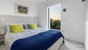 A bedroom with a bed, a painting, a nightstand, a window, and a balcony.