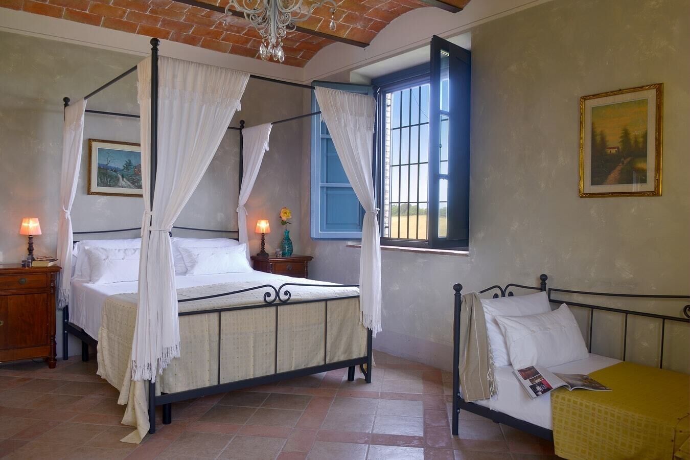 Ein Zimmer mit Himmelbett, einem zusätzlichen Bett, einem Nachttisch mit Lampe, einem Fenster mit Vorhängen und einem Bild an der Wand.