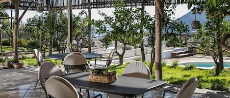 An outdoor patio with tables and chairs, surrounded by greenery and a pool.