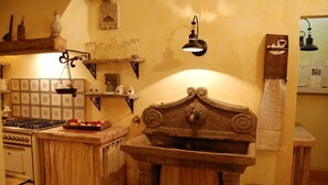 Een keuken met een stenen spoelbak, houten planken met glazen potten, een fornuis en een hangende lamp.