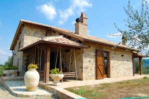 Une maison en pierre avec un toit en tuiles, une cheminée et un porche doté d’une balançoire.