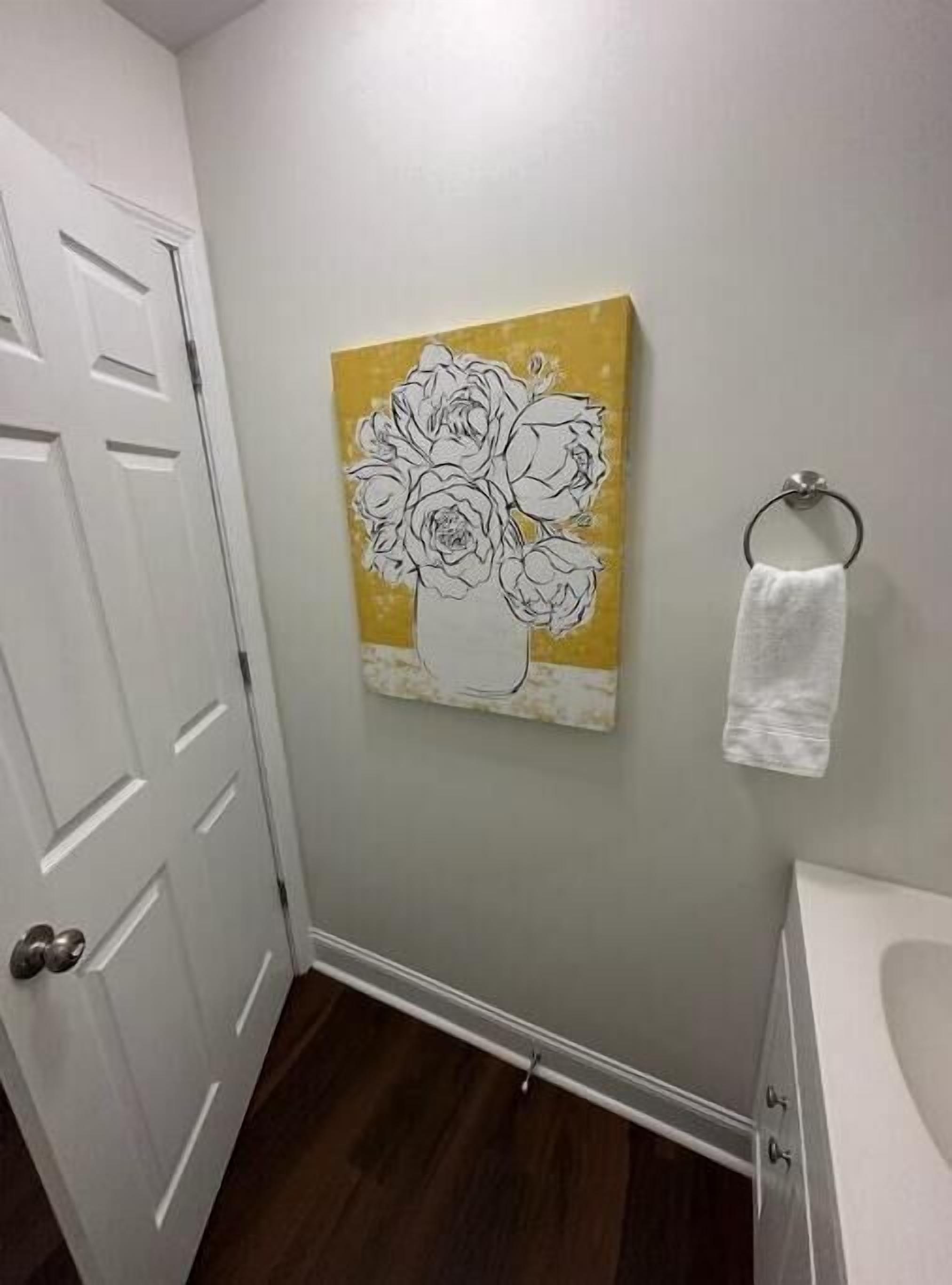 Un baño con una puerta blanca, una toalla blanca colgada en un aro metálico y una obra de arte en la pared que muestra un jarrón con flores.