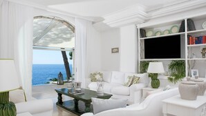 Un salon décoré dans des tons de blanc, avec vue sur la mer, une bibliothèque et un téléviseur.