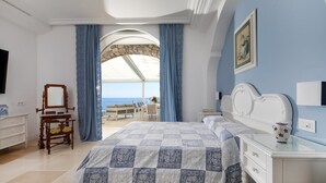 Une chambre avec un grand lit, une coiffeuse, une commode et une vue sur la mer, visible à travers une arche.