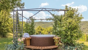 Ein Whirlpool mit einer Holzterrasse und einer Metall-Pergola, umgeben von üppiger Vegetation und einer Bergkulisse.