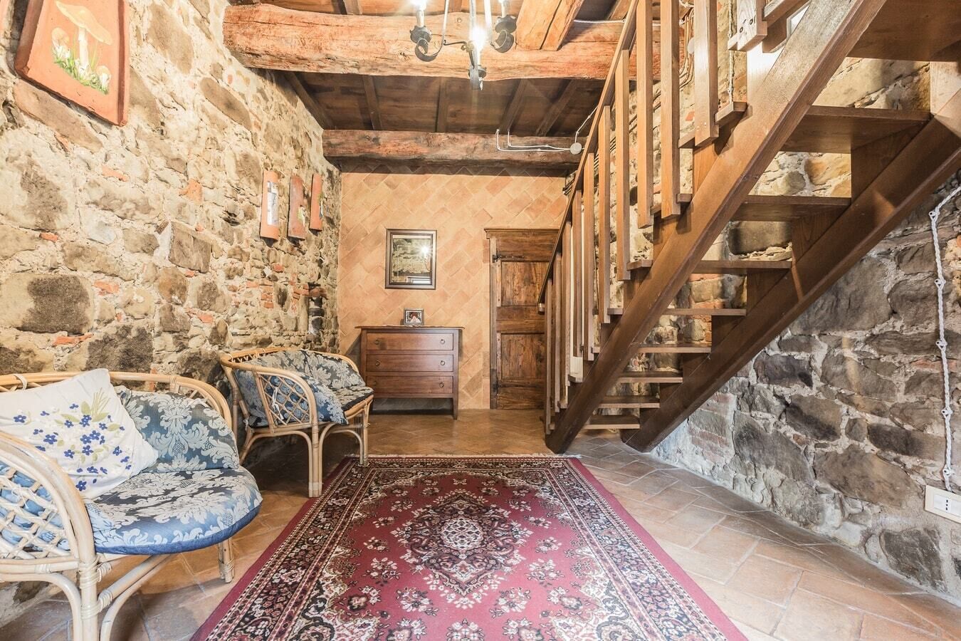 A rustic interior with stone walls, wooden beams, a staircase, and a patterned rug.