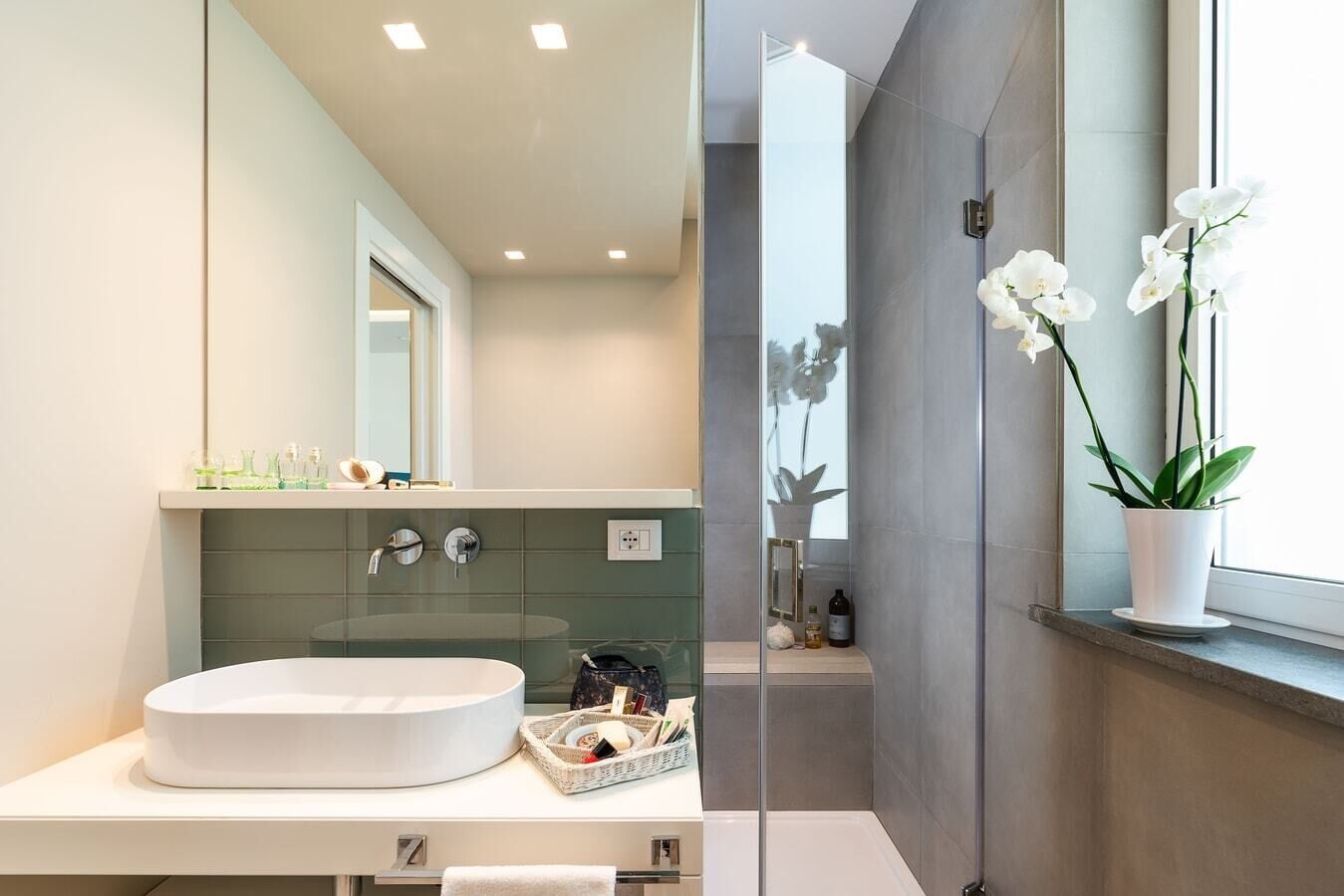 A modern bathroom with a large oval sink, a glass shower enclosure, and a potted orchid on the windowsill.